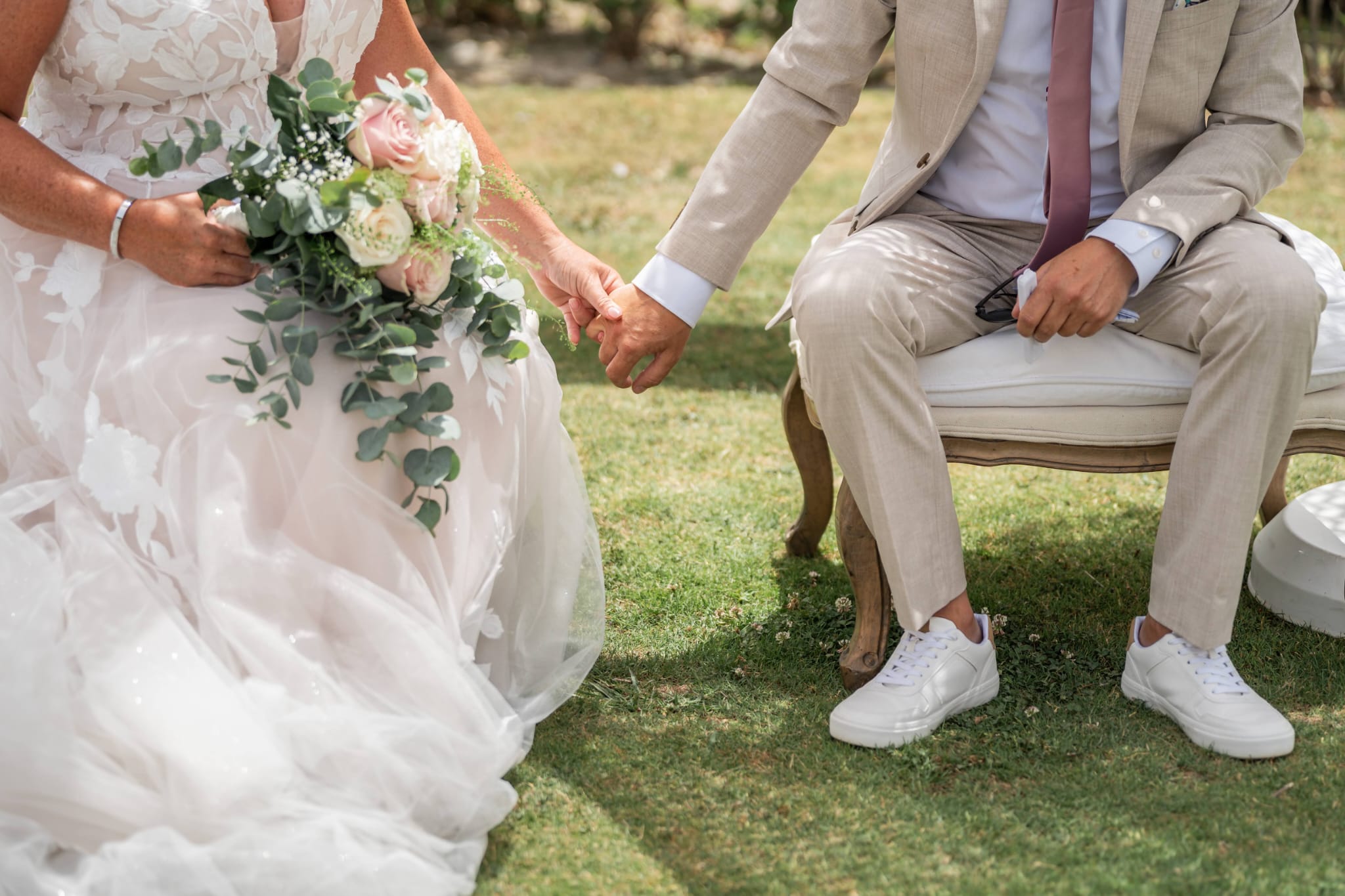 Bride and groom holding hands at an outdoor ceremony, her bouquet of blush roses and eucalyptus in her lap